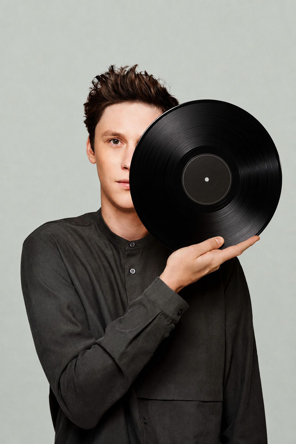 Portrait of FLYIN HIAS holding a vinyl record that covers half of the face, light studio backdrop